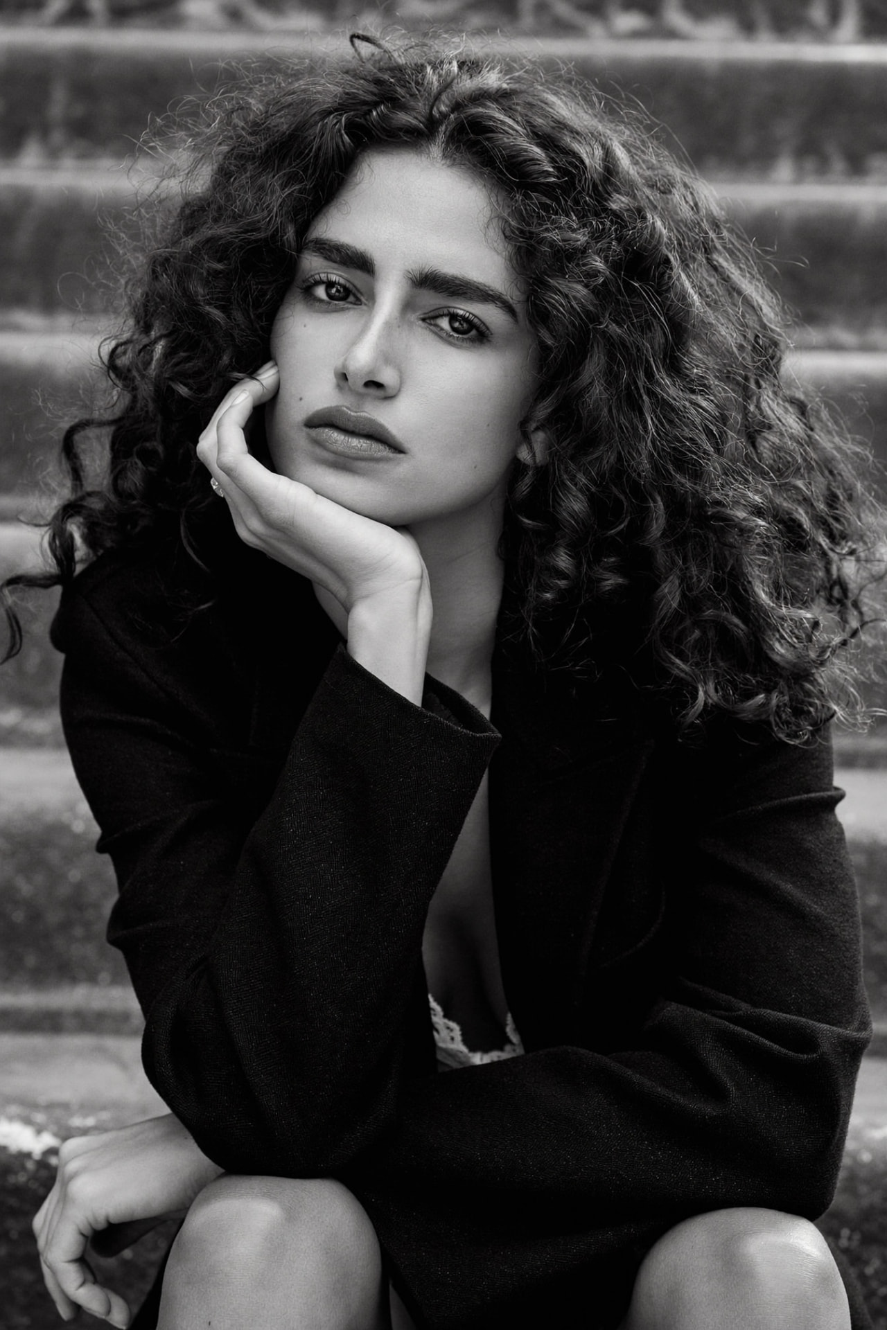 Black-and-white portrait of a curly-haired model resting her chin on her hand on stone steps
